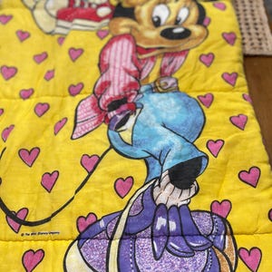May include: A yellow sleeping bag featuring a cartoon character with a large head, wearing a blue hat and purple purse. The sleeping bag is decorated with pink hearts and the text "The Walt Disney Company."