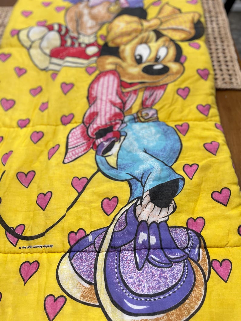 May include: A yellow sleeping bag featuring a cartoon character with a large head, wearing a blue hat and purple purse. The sleeping bag is decorated with pink hearts and the text "The Walt Disney Company."
