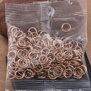 May include: A clear plastic bag filled with a large quantity of small, gold-colored metal jump rings. The jump rings are all the same size and are arranged in a random pattern.