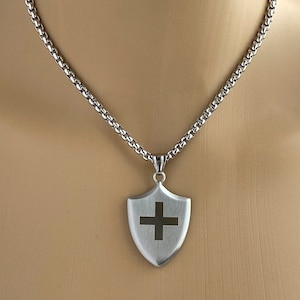May include: A silver chain necklace with a silver shield pendant featuring a black cross.
