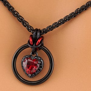 May include: Black metal necklace with a heart-shaped pendant. The pendant has a large, faceted red gemstone and a black stone border. The necklace has a black chain and a black ring that holds the pendant.