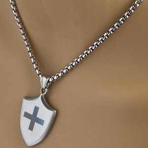 May include: A silver chain necklace with a silver shield pendant featuring a black cross. The shield is slightly textured.