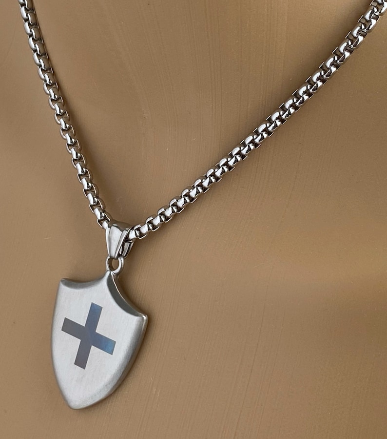 May include: A silver chain necklace with a silver shield pendant featuring a black cross. The shield is slightly textured.