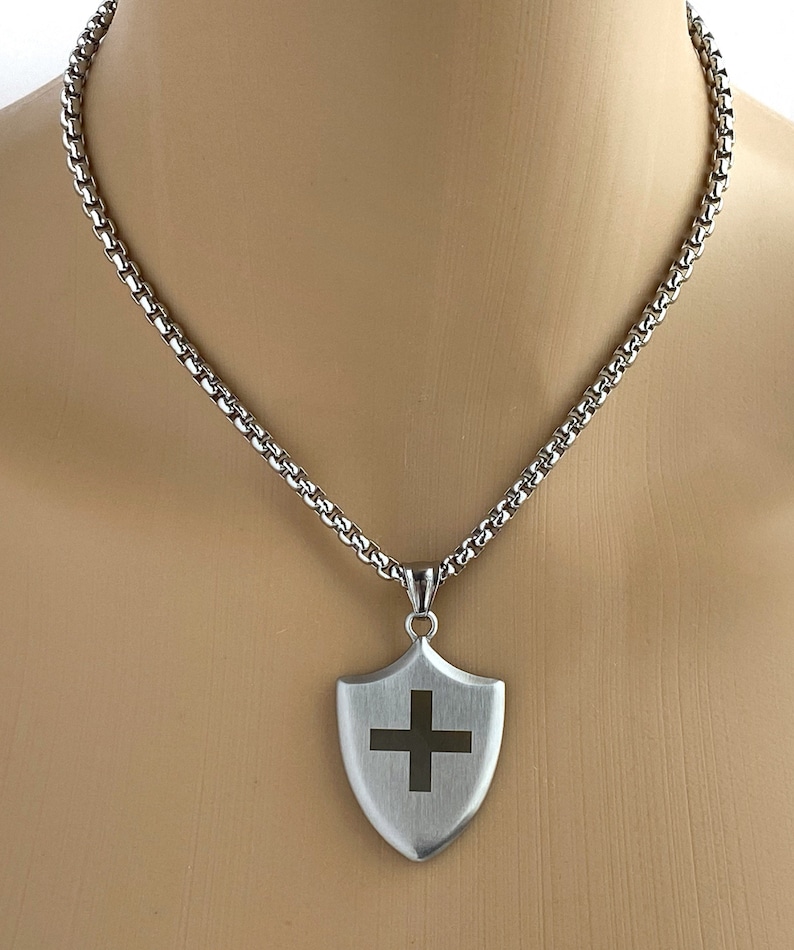 May include: A silver chain necklace with a silver shield pendant featuring a black cross.