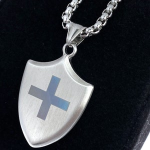 May include: A silver chain necklace with a silver shield pendant featuring a blue cross. The pendant has a brushed metal finish.