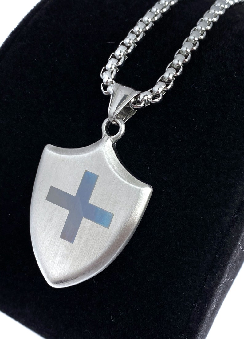 May include: A silver chain necklace with a silver shield pendant featuring a blue cross. The pendant has a brushed metal finish.