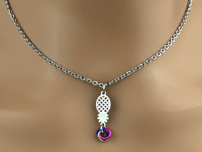 Puede incluir: Un collar de cadena plateada con un colgante de pi&ntilde;a y un conjunto de tres anillos entrelazados en morado, rosa y plateado.