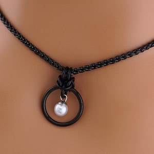 K&ouml;nnte beinhalten: Schwarze Halskette mit einem kreisf&ouml;rmigen Anh&auml;nger. Der Anh&auml;nger hat einen schwarzen Ring mit einer kleinen, wei&szlig;en Perle in der Mitte. Die Halskette hat ein gewebtes Kettendesign.