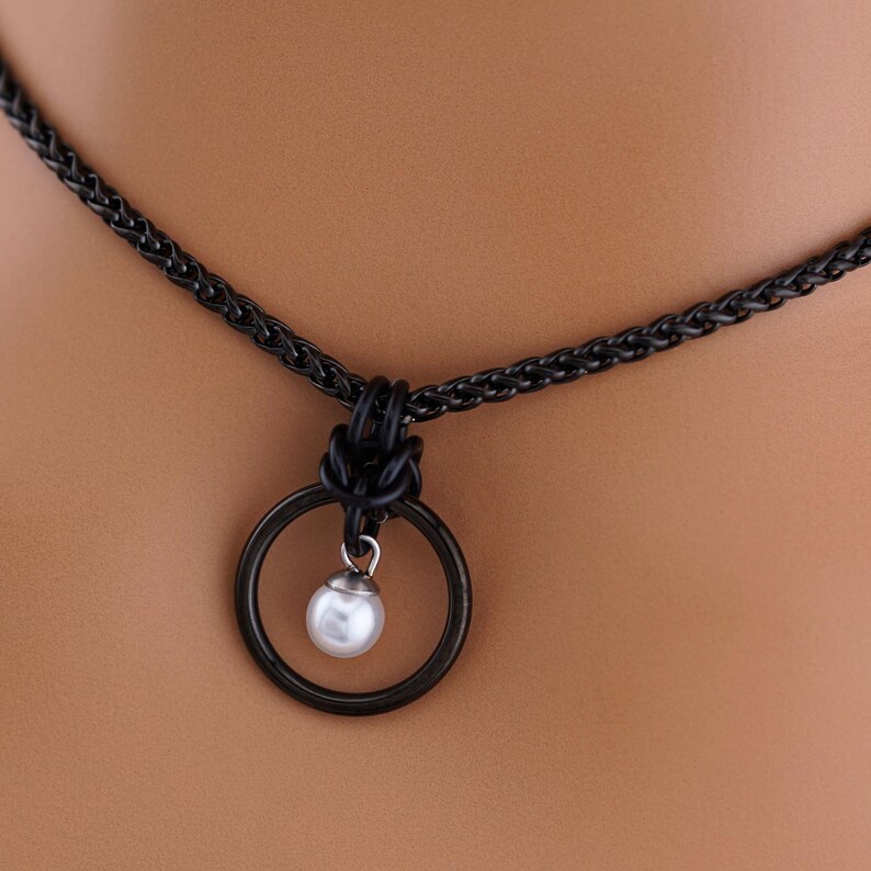 K&ouml;nnte beinhalten: Schwarze Halskette mit einem kreisf&ouml;rmigen Anh&auml;nger. Der Anh&auml;nger hat einen schwarzen Ring mit einer kleinen, wei&szlig;en Perle in der Mitte. Die Halskette hat ein gewebtes Kettendesign.