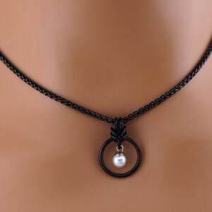 K&ouml;nnte beinhalten: Eine schwarze Halskette mit einem kreisf&ouml;rmigen Anh&auml;nger und einer kleinen, h&auml;ngenden wei&szlig;en Perle. Die Kette hat eine geflochtene Kette und ein dunkles Finish, wodurch ein minimalistisches Design entsteht. Die Perle ist rund und hat einen subtilen Glanz.