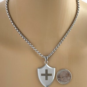 May include: A silver chain necklace with a silver shield pendant featuring a cross. The pendant is about the size of a US quarter.