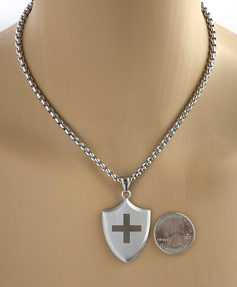 May include: A silver chain necklace with a silver shield pendant featuring a cross. The pendant is about the size of a US quarter.