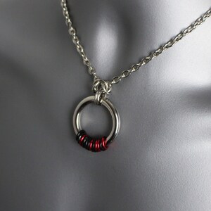 May include: A silver chain necklace with a silver ring pendant. The ring is decorated with black and red wire wrapped around it.