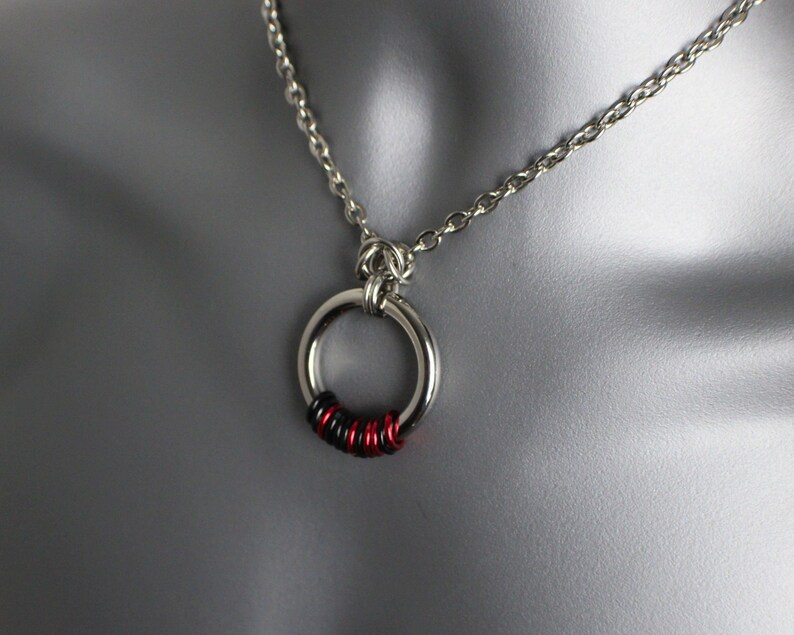 May include: A silver chain necklace with a silver ring pendant. The ring is decorated with black and red wire wrapped around it.