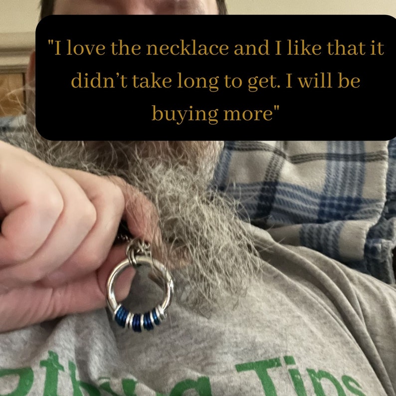May include: A close-up of a silver necklace with a blue and silver ring pendant. The necklace is worn by a person with a long grey beard. The text "I love the necklace and I like that it didn't take long to get. I will be buying more" is visible in the image.