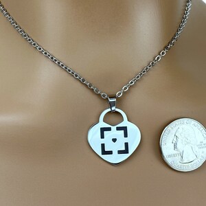 May include: A silver heart-shaped pendant necklace with a black square design and a small black heart in the center. The necklace is on a silver chain and is shown on a model's neck.
