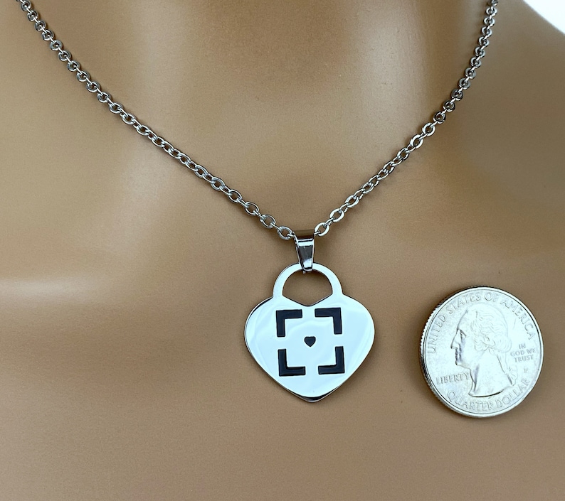 May include: A silver heart-shaped pendant necklace with a black square design and a small black heart in the center. The necklace is on a silver chain and is shown on a model's neck.