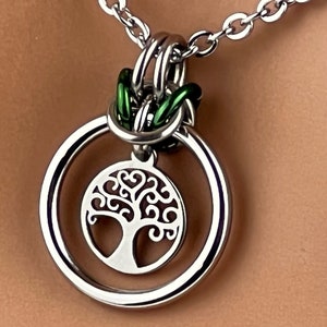 May include: A silver necklace with a green chain and a pendant featuring a tree of life design. The pendant is a double circle with the tree of life design in the center.