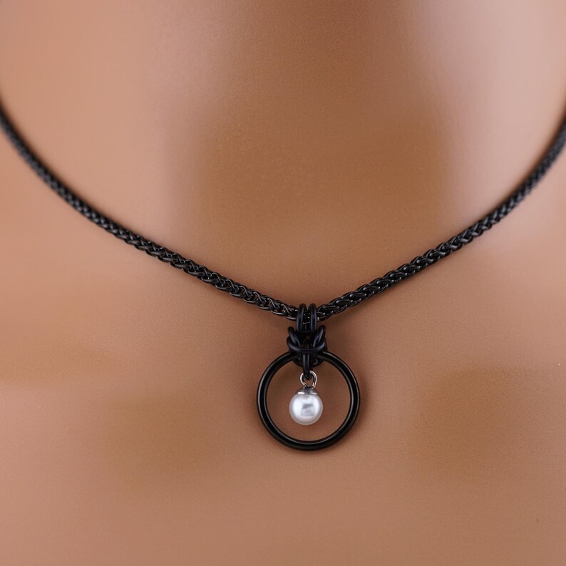 K&ouml;nnte beinhalten: Eine schwarze Halskette mit einem kreisf&ouml;rmigen Anh&auml;nger. Der Anh&auml;nger hat eine kleine, wei&szlig;e Perle, die in einem schwarzen Ring h&auml;ngt. Die Halskette ist ebenfalls schwarz.