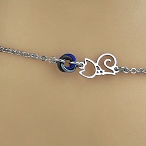 May include: A silver chain bracelet with a small cat charm and a multi-colored ring charm. The ring charm is made of blue, gray, and white.