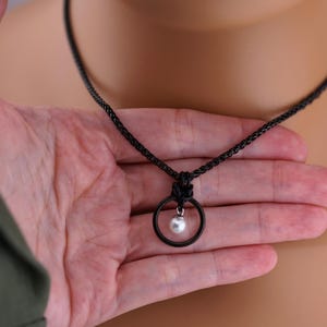 K&ouml;nnte beinhalten: Eine schwarze Halskette mit einem kreisf&ouml;rmigen Anh&auml;nger mit einer kleinen, wei&szlig;en Perle. Der Anh&auml;nger h&auml;ngt an einer schwarzen Kette. Die Halskette wird in einer Hand gehalten, um das Design und die Details des Schmucks hervorzuheben.