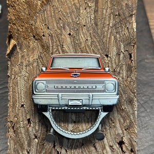 May include: A silver bottle opener shaped like a vintage red Chevrolet pickup truck. The truck has a silver grill and chrome bumpers. The bottle opener is mounted on a wooden surface.