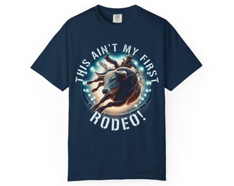 May include: Navy blue t-shirt featuring a rodeo design. The graphic depicts a bull rider on a bucking bull, with the text "THIS AIN'T MY FIRST RODEO!" in white. The design is set against a backdrop of stadium lights.