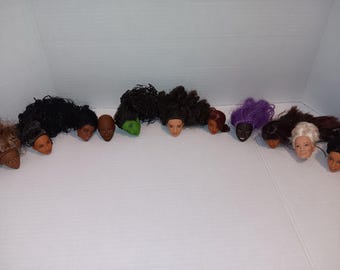 May include: A collection of eleven doll heads with diverse skin tones and hair styles, including long black curly hair, short brown hair, a green-skinned head, purple hair, and white hair, arranged in a horizontal row against a plain background.