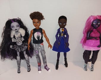 May include: Four fashion dolls standing in a row. The first has long black hair, a white fur collar, and a striped top. The second has short curly hair, a black graphic tank top, and gray pants. The third wears a blue striped blazer and skirt. The fourth has long pink hair and a pink outfit.