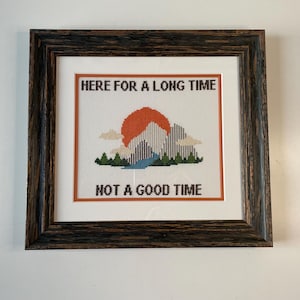 May include: Framed cross-stitch art with a mountain and sunset design. The text reads "HERE FOR A LONG TIME NOT A GOOD TIME". The frame is dark brown with a textured appearance. The artwork is on a white background.