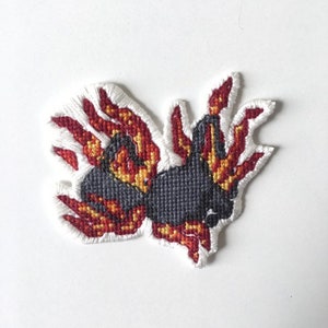 May include: A black and red embroidered patch of a creature with flames for wings. The creature is in a flying pose with its wings spread out.