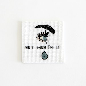 May include: White cross-stitch embroidery with a black eye, a single teardrop, and the text "NOT WORTH IT".