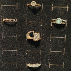 Könnte beinhalten: Eine Sammlung von Ringen, die auf einer schwarzen Oberfläche präsentiert werden. Die Ringe weisen verschiedene Designs auf, darunter ein verdrehtes Band mit einem klaren Stein, ein Band mit kleinen Steinen und ein goldener Ring mit einer einzigartigen geometrischen Form und einem klaren Stein.