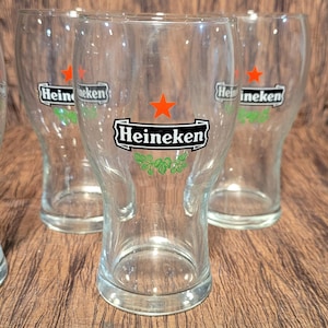 May include: Clear glass beer glasses with the Heineken logo. The logo features a red star, the word "Heineken" in a black rectangle, and green hops. The glasses are on a wooden surface.
