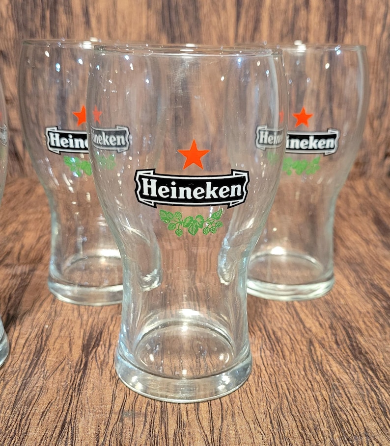 May include: Clear glass beer glasses with the Heineken logo. The logo features a red star, the word "Heineken" in a black rectangle, and green hops. The glasses are on a wooden surface.