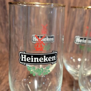 May include: Clear glass Heineken beer glasses with gold rims. The glasses feature the Heineken logo in black and white with a red windmill and green hops design. The glasses are empty and appear to be clean.
