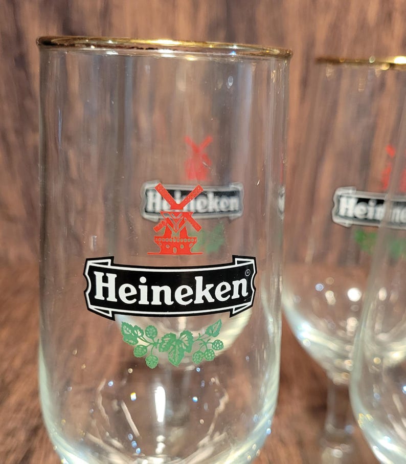 May include: Clear glass Heineken beer glasses with gold rims. The glasses feature the Heineken logo in black and white with a red windmill and green hops design. The glasses are empty and appear to be clean.