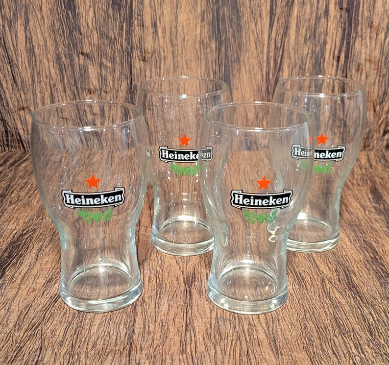May include: Four clear glass beer glasses with the Heineken logo. Each glass has a curved shape and a wide mouth. The logo features a red star and green accents. The glasses are arranged on a brown surface.