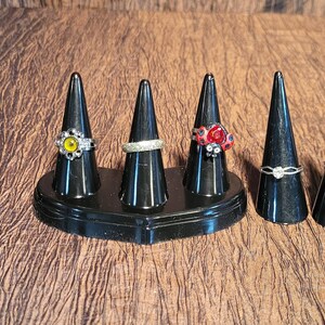 May include: Six rings displayed on black cones. The rings have diverse designs, including a yellow stone, a band of clear stones, and a sunburst design. The cones are on a brown, textured surface.