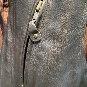 May include: Close-up of a black leather jacket with a zipped pocket. The zip is metallic and has a leather pull tab. The leather has a textured appearance.