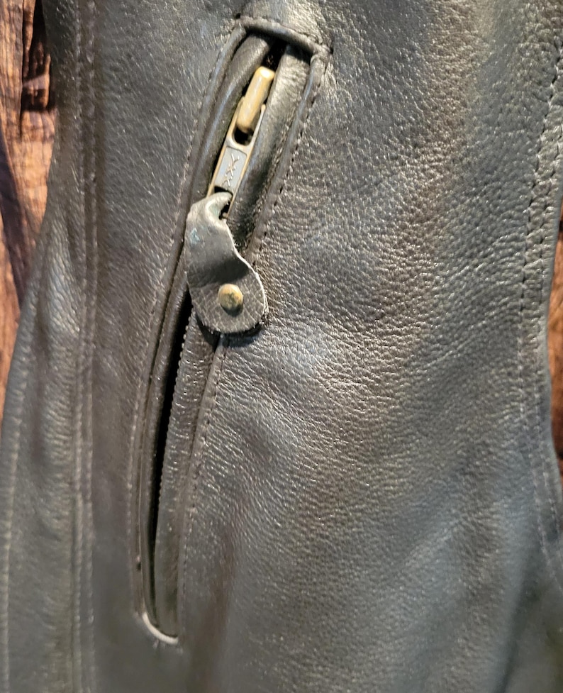 May include: Close-up of a black leather jacket with a zipped pocket. The zip is metallic and has a leather pull tab. The leather has a textured appearance.