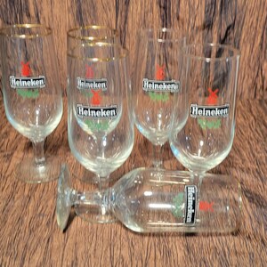 May include: A collection of clear, stemmed beer glasses with gold rims. Each glass features the "Heineken" logo in black, red, and green. One glass is lying on its side. The glasses are arranged on a brown surface.