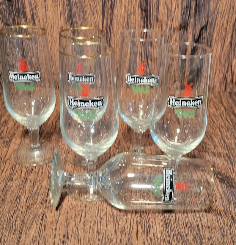 May include: A collection of clear, stemmed beer glasses with gold rims. Each glass features the "Heineken" logo in black, red, and green. One glass is lying on its side. The glasses are arranged on a brown surface.