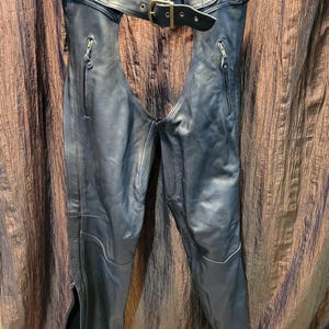 May include: Black leather motorcycle chaps with a belt and buckle closure. The chaps have zippered pockets and zippers along the outer leg seams. The top of the chaps has the letters "LK" on the inside.
