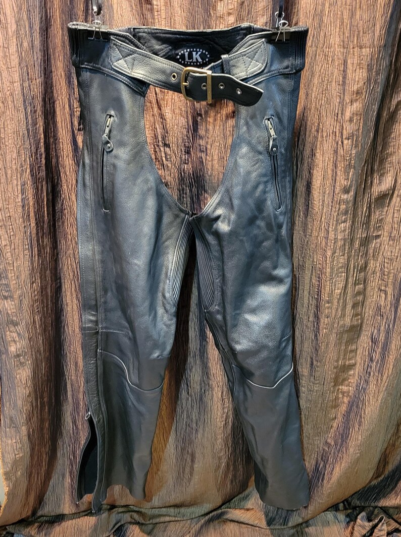 May include: Black leather motorcycle chaps with a belt and buckle closure. The chaps have zippered pockets and zippers along the outer leg seams. The top of the chaps has the letters "LK" on the inside.