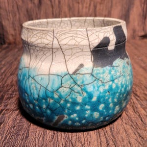 May include: A handcrafted ceramic bowl with a unique crackle glaze. The bowl features a gradient of colors, transitioning from white to turquoise and dark blue. The rim has a black accent.