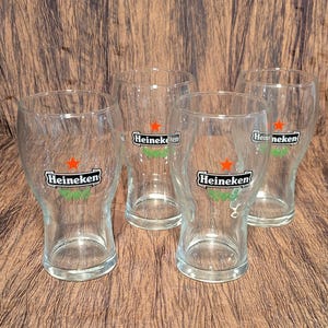 May include: Four clear glass beer glasses with the Heineken logo, featuring a red star and green accents. The glasses are empty and have a tulip shape, ideal for serving beer. The glasses are arranged on a brown surface.