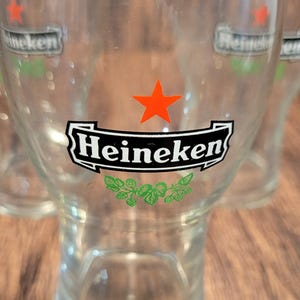 May include: Clear glass beer glasses with the Heineken logo. The logo features a black banner with the word "Heineken" in white, a red star, and green accents. The glasses are on a wooden surface.