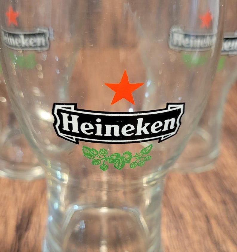 May include: Clear glass beer glasses with the Heineken logo. The logo features a black banner with the word "Heineken" in white, a red star, and green accents. The glasses are on a wooden surface.