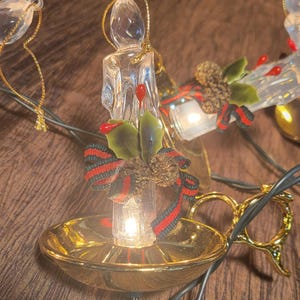 May include: A close-up of a Christmas light string featuring clear, candle-shaped lights with gold-colored bases. Each candle is adorned with holly, red berries, and a red and green ribbon. The lights are on, illuminating the details.